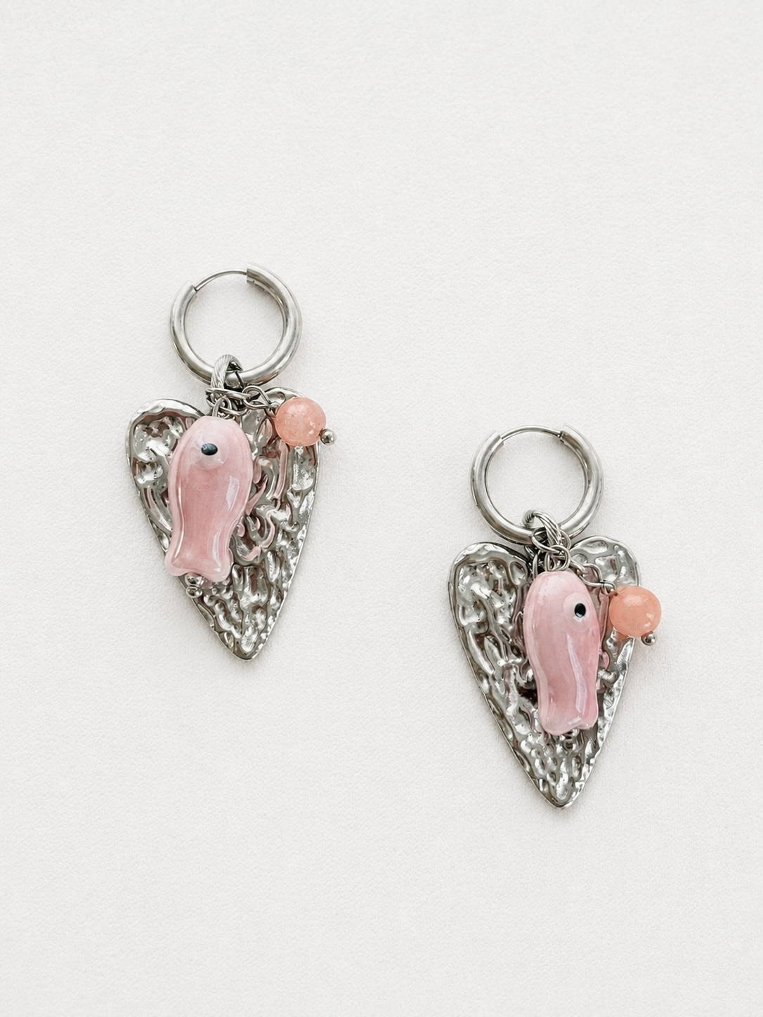 Hnadgemaakte zilveren RVS oorbellen met statement hart en roze vis aan een 12 mm oorring